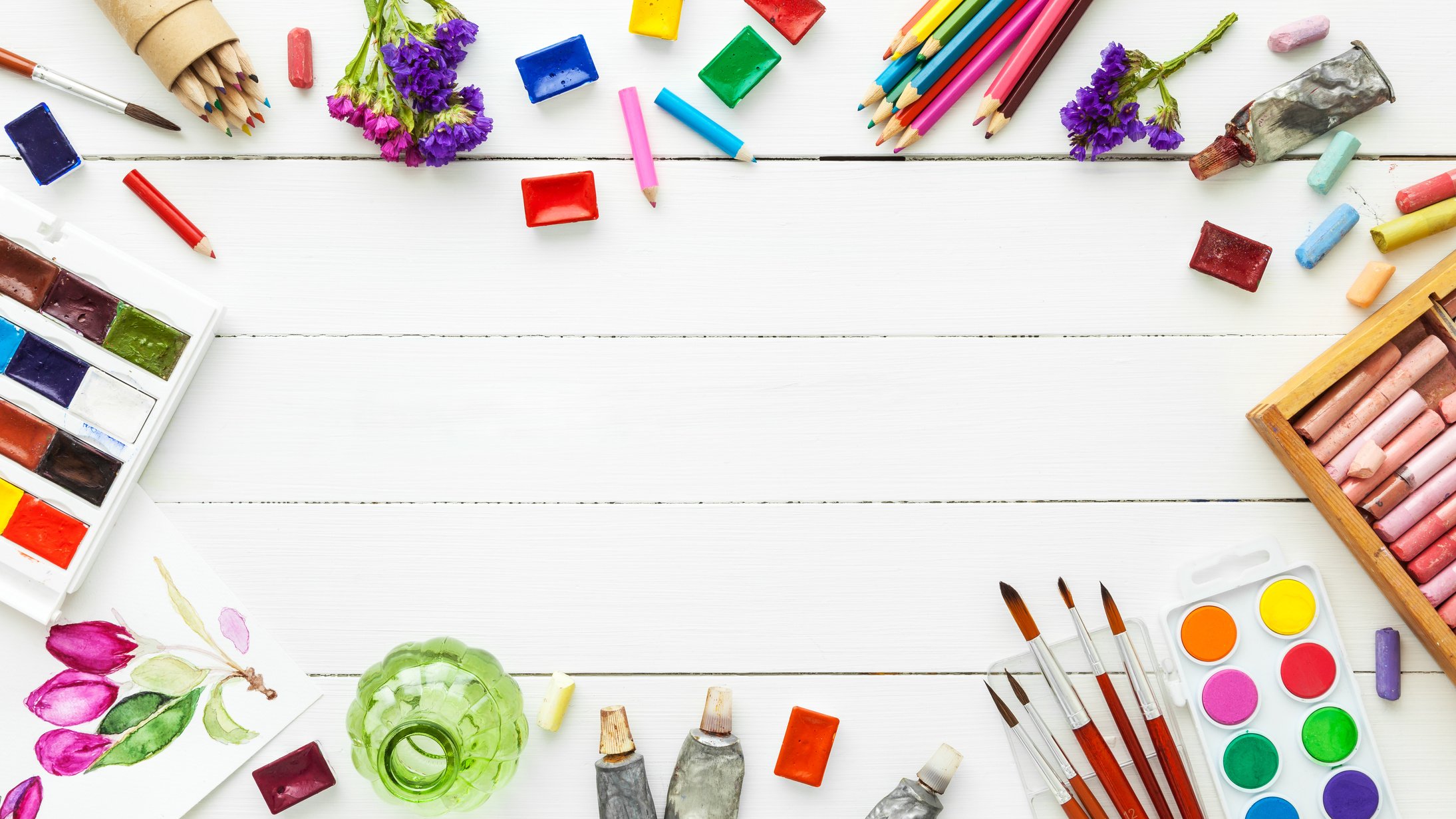 Watercolor paints, brushes, pencils, pastel crayon on white table.